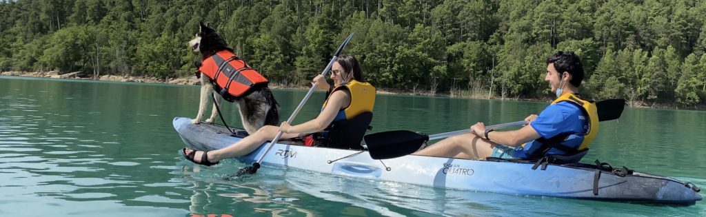 Un perro apoyando sus dos patas delanteras en la parte delantera de un kayak y una mujer y un hombre detrás, sujetando los remos.
